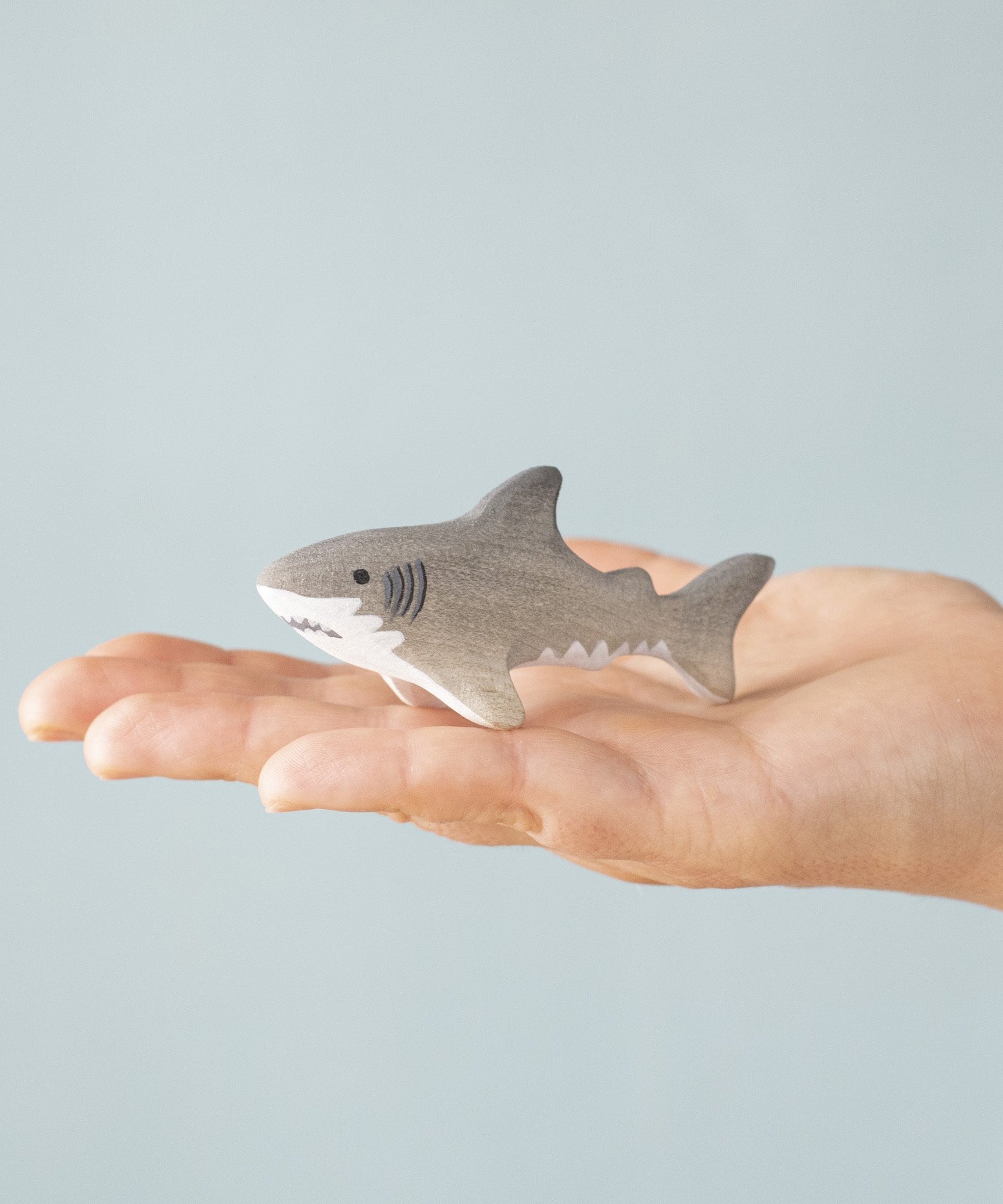 An adult hand holding the Bumbu handmade and hand painted wooden grey baby shark figure showing size