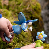 Adult hand holding Bumbu's wooden blue forget me not flower child figure next to a blue fairy figure on moss