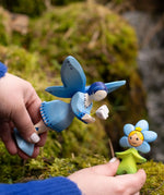 Adult hand holding Bumbu's wooden blue forget me not flower child figure next to a blue fairy figure on moss