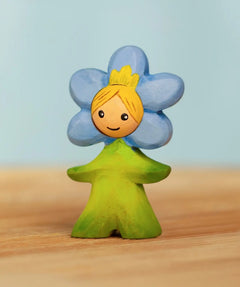 Bumbu handcrafted wooden blue forget me not flower child figure with hand painted facial features standing on a wooden surface