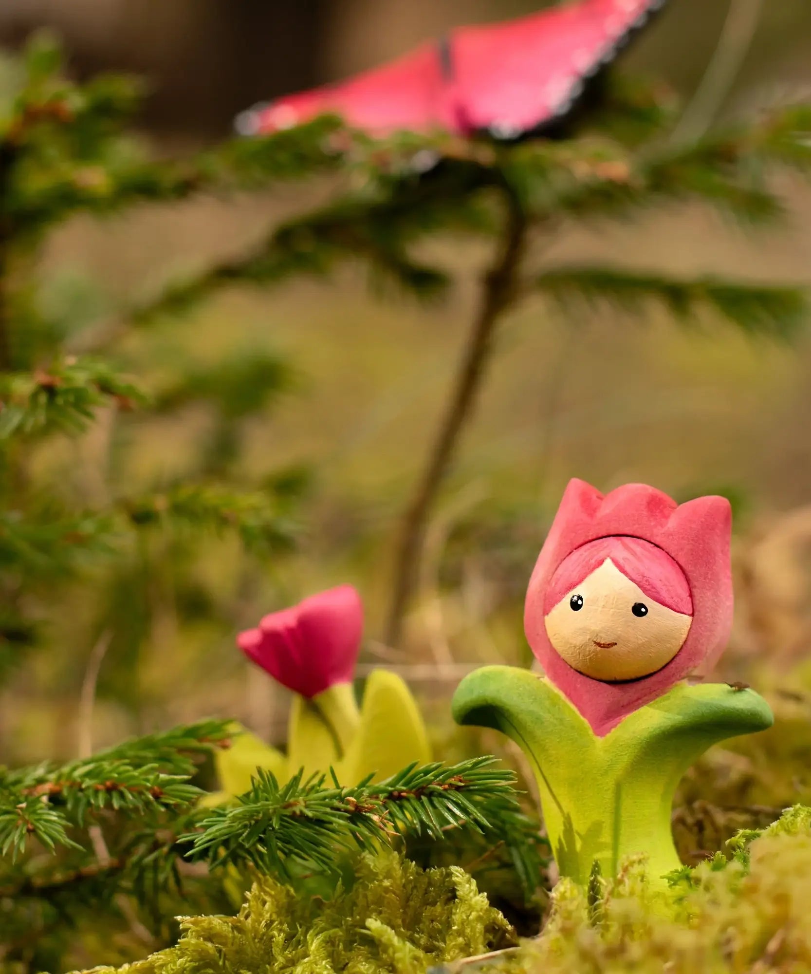 Bumbu wooden pink tulip figure showing hand painted facial features standing in moss with a butterfly in the background.