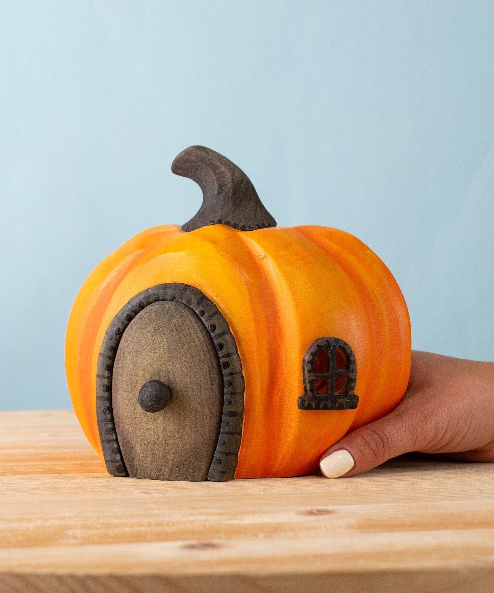 An adult hand holding the Bumbu handmade and hand painted wooden halloween orange pumpkin house showing scale