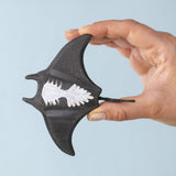 An adult hand holding the Bumbu handmade and hand painted wooden black manta ray figure showing the underside detail