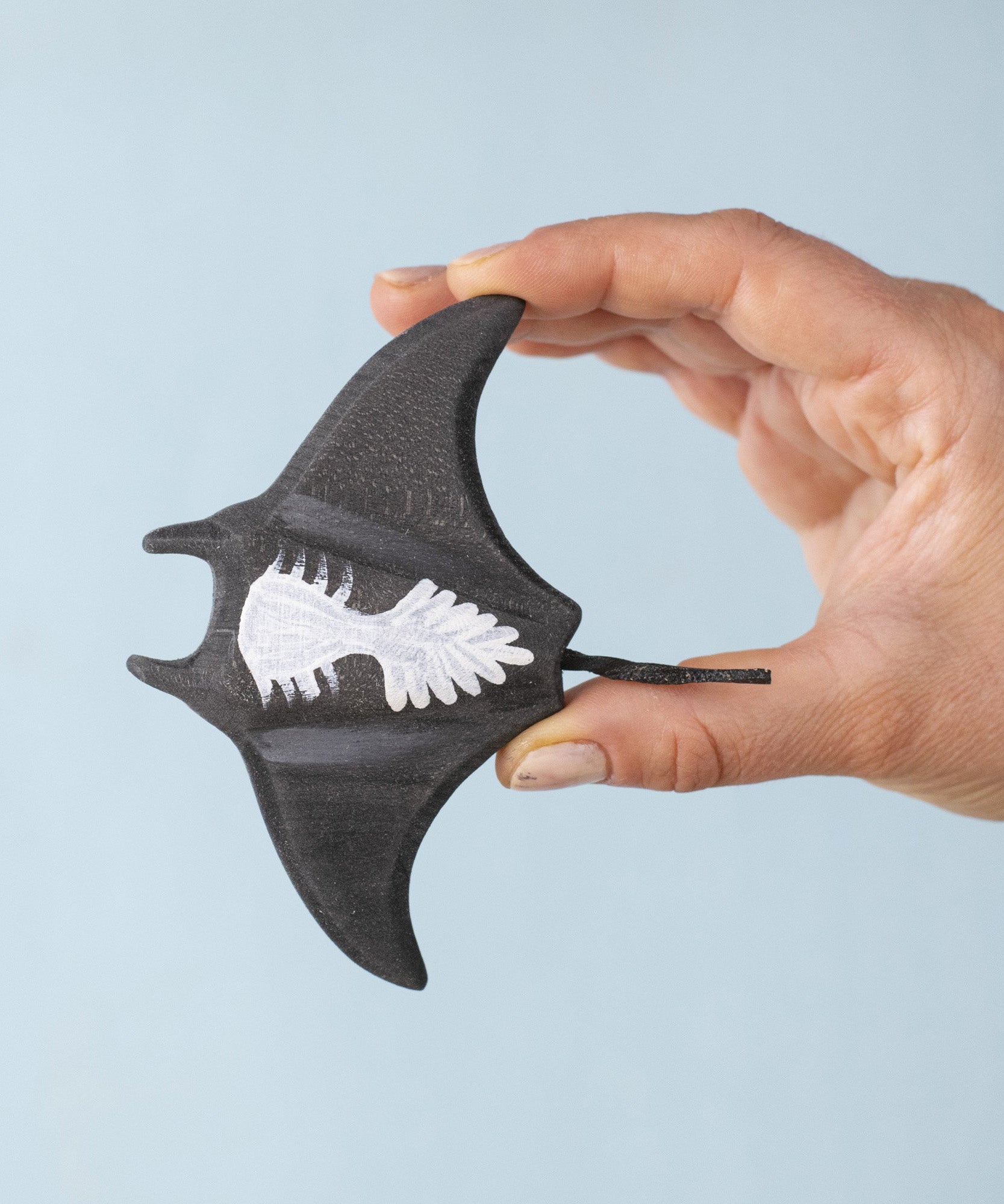 An adult hand holding the Bumbu handmade and hand painted wooden black manta ray figure showing the underside detail