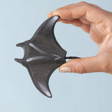 An adult hand holding the Bumbu handmade and hand painted wooden black manta ray figure showing the topside detail