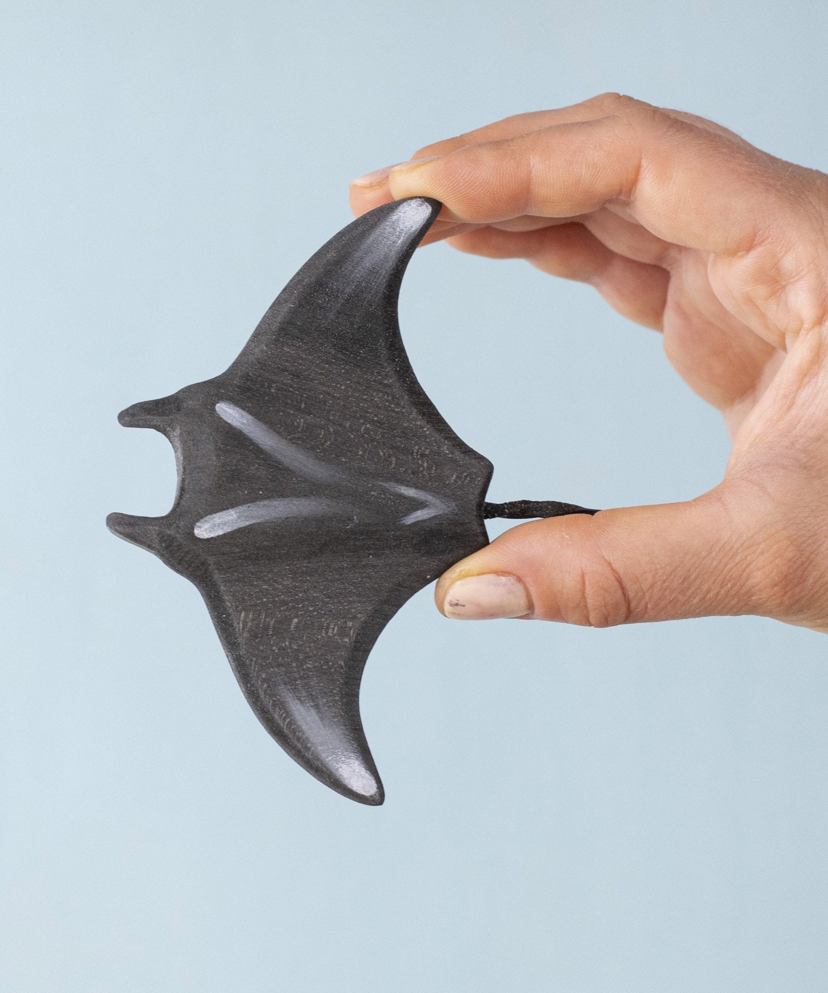 An adult hand holding the Bumbu handmade and hand painted wooden black manta ray figure showing the topside detail