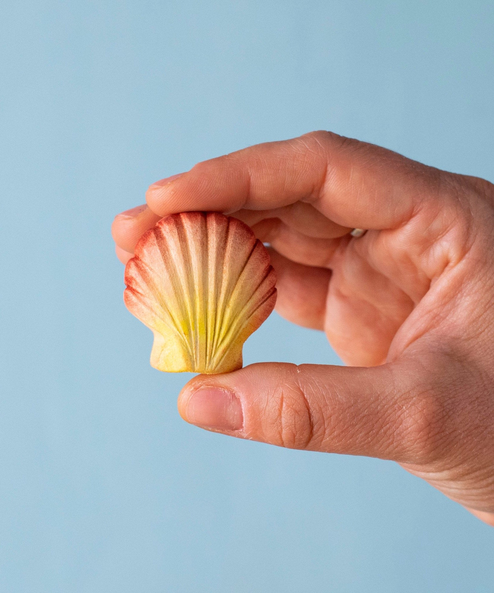 An adult hand holding the Bumbu handmade and hand painted orange and red wooden sea shell figure showing size