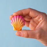 An adult hand holding the Bumbu handmade and hand painted orange and pink wooden sea shell toy figure showing size