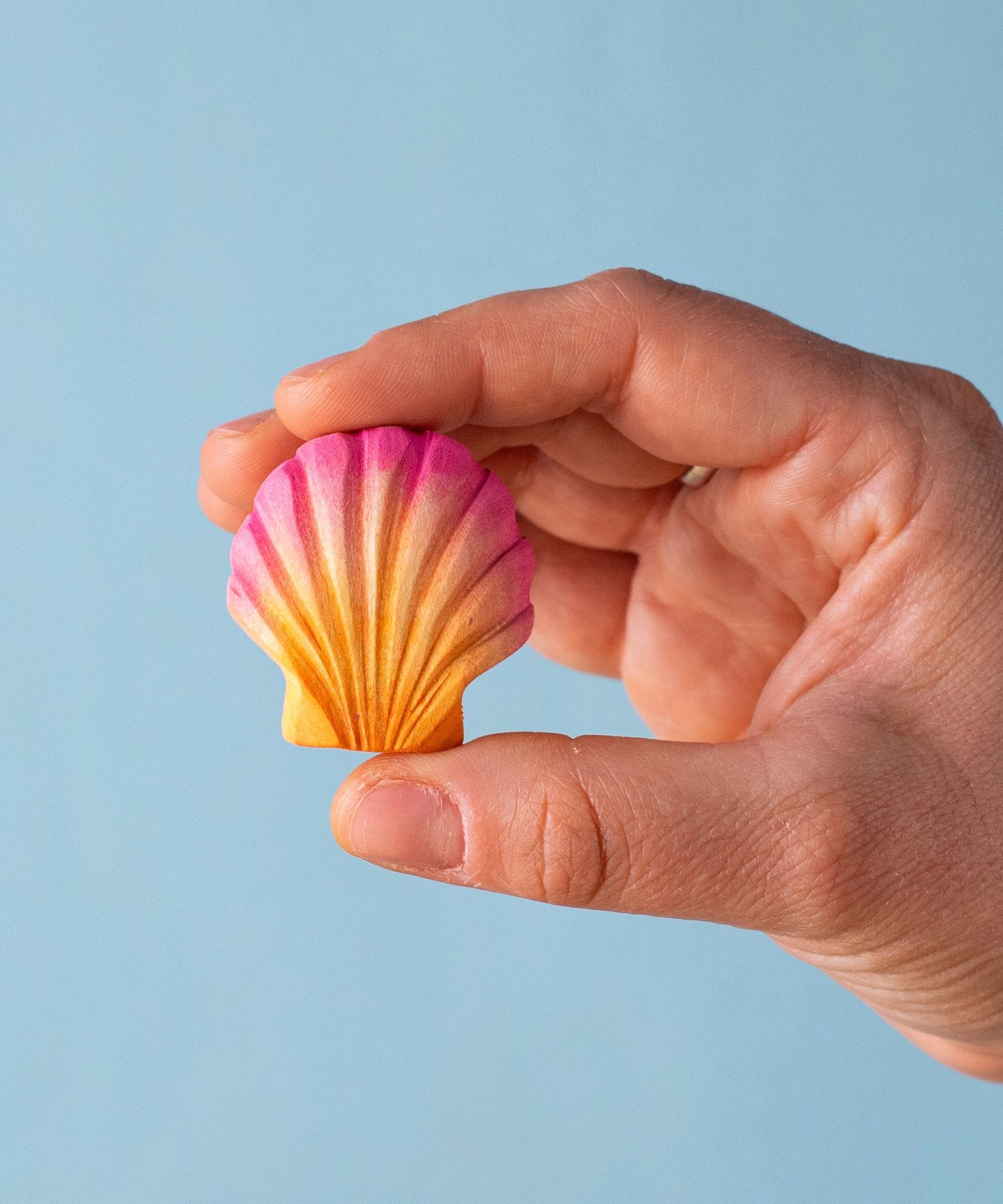 An adult hand holding the Bumbu handmade and hand painted orange and pink wooden sea shell toy figure showing size