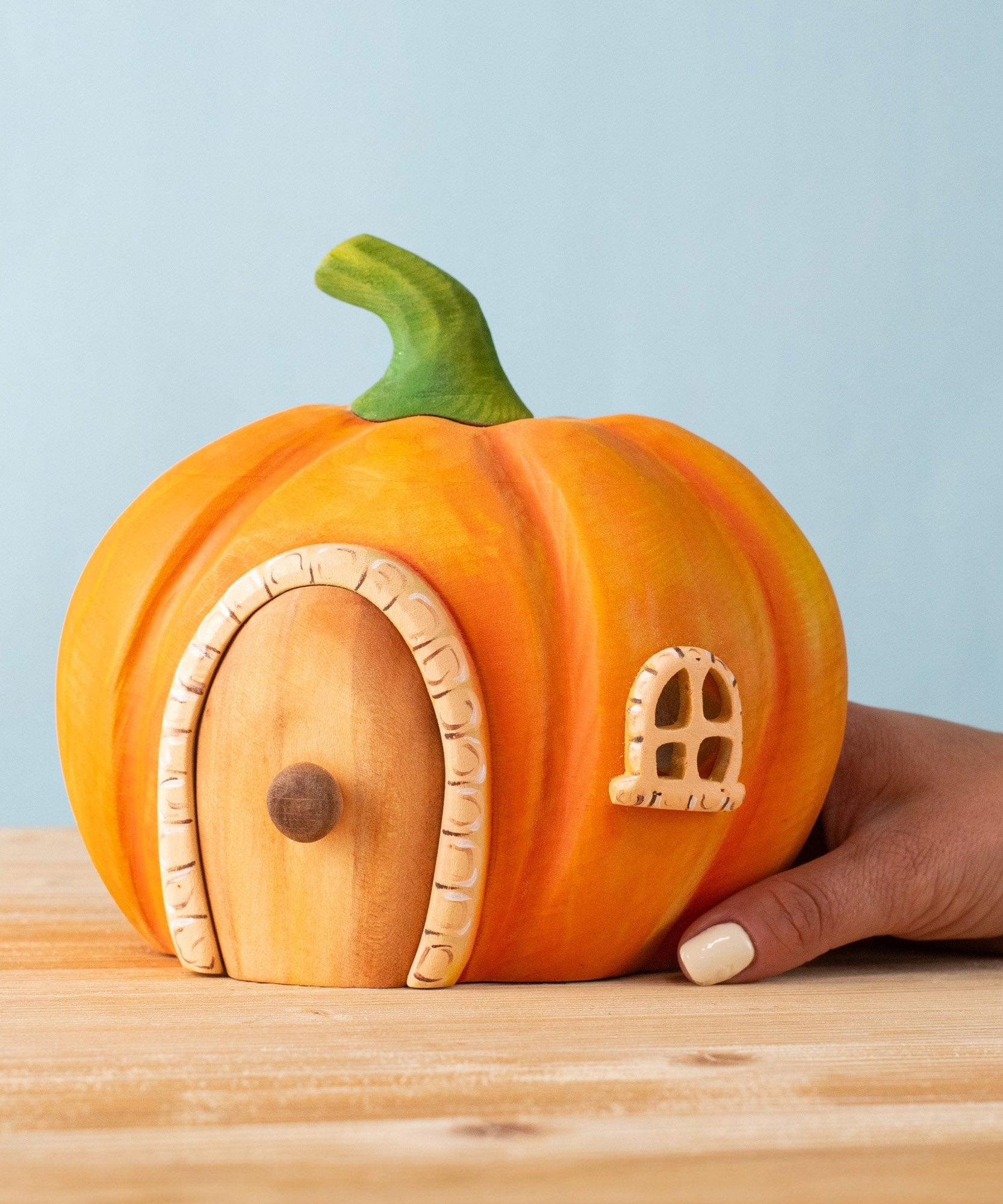 An adult hand holding the Bumbu handmade and hand painted wooden orange pumpkin house showing scale