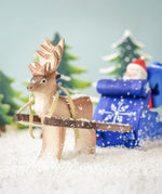 A snowy scene of a Bumbu Reindeer pulling the Bumbu Santa in the Blue Snow Sleigh. In the background are snowy green Bumbu Trees