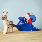 Bumbu Santa sitting in the back of the Snow Sleigh with a blue wooden sack on his back, and a Reindeer attached to the reins of the sleigh