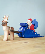 Bumbu Santa sitting in the back of the Snow Sleigh with a blue wooden sack on his back, and a Reindeer attached to the reins of the sleigh