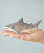 An adult hand holding the Bumbu handmade and hand painted wooden grey shark figure showing size