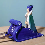 The Ice Queen is sitting in the back of the blue Snow Sleigh. The image shows the sleigh from the side where you can see the decorative white painted designs and the brown reins are attached at the front of the sleigh, slightly showing
