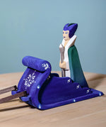 The Ice Queen is sitting in the back of the blue Snow Sleigh. The image shows the sleigh from the side where you can see the decorative white painted designs and the brown reins are attached at the front of the sleigh, slightly showing