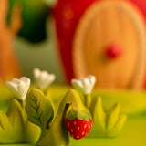 Bumbu handcrafted wooden strawberry plant figure next to white wooden flowers and a strawberry house in the background.