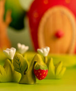 Bumbu handcrafted wooden strawberry plant figure next to white wooden flowers and a strawberry house in the background.