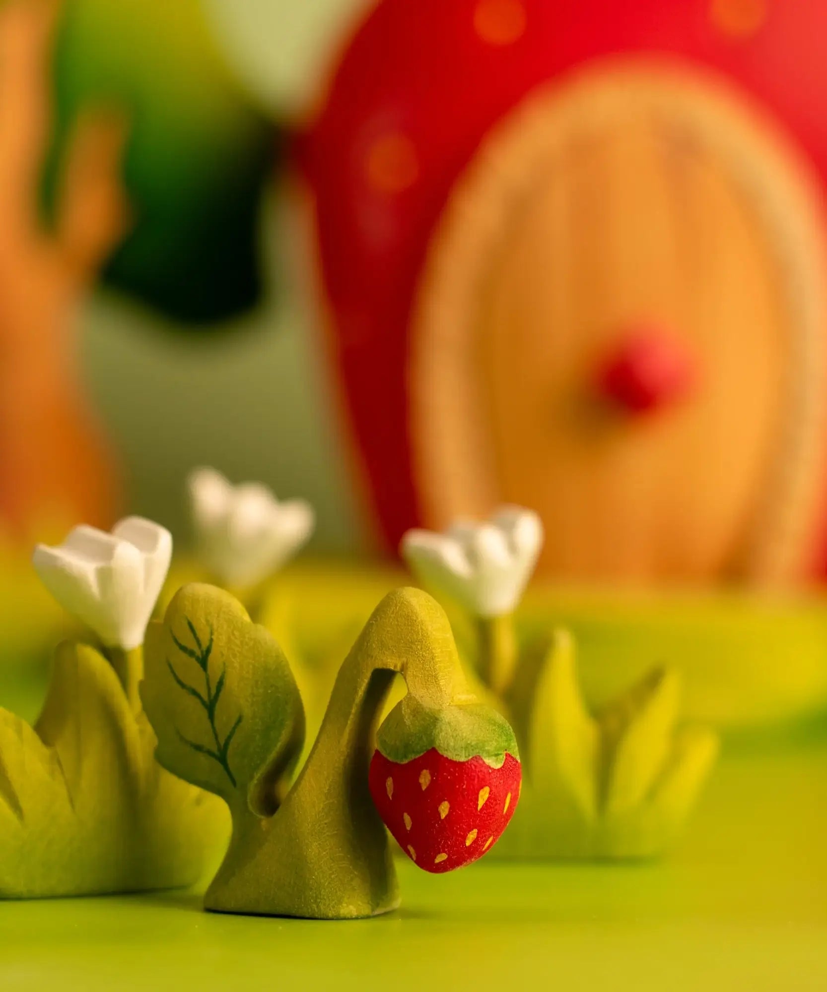 Bumbu handcrafted wooden strawberry plant figure next to white wooden flowers and a strawberry house in the background.