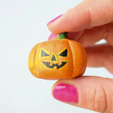 The small Bumbu Toys Halloween Pumpkin is being held in between a persons fingers, they are wearing pink nail polish. This image shows the detailed facial features and pumpkin grooves in the woodwork