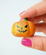 The small Bumbu Toys Halloween Pumpkin is being held in between a persons fingers, they are wearing pink nail polish. This image shows the detailed facial features and pumpkin grooves in the woodwork