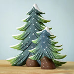 The Bumbu Toys Large and Small wooden snowy fir trees together, stood on a wooden table with a light blue background The large fir tree is behind the small fir tree