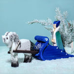 The Snow Queen riding on the Bumbu Wooden Snow Sleigh, with a wooden white horse attached to the reins of the sleigh. The background is a snowy landscape with a snowy green tree