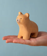 An adult hand holding the Bumbu handmade and hand painted wooden brown curious bear figure showing