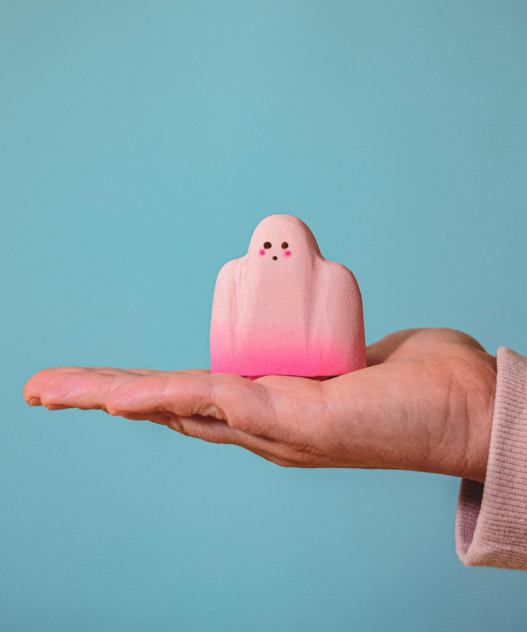 An adult hand holding the Bumbu handmade and hand painted wooden pink ghost figure for size reference