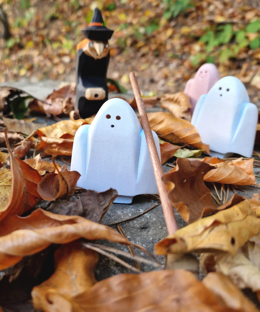 A group of white and pink Bumbu wooden ghost figures and a witch figure standing on autumn leaves