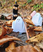 A group of white and pink Bumbu wooden ghost figures and a witch figure standing on autumn leaves