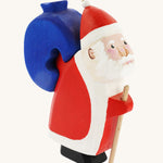 Bumbu Wooden Santa Claus, is a handmade and hand painted festive toy figure with a wooden blue sack that can be removed from its back. The image shows Santa with rosy cheeks, a beard, a red and white outfit and a red and white hat, with the blue wooden sack on his back. The image is on a cream background