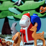 The Bumbu Wooden Santa toy is in the woods with woodland animals which include a Bumbu rabbit, a Bumbu deer and green trees in the background