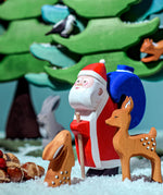 The Bumbu Wooden Santa toy is in the woods with woodland animals which include a Bumbu rabbit, a Bumbu deer and green trees in the background