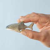 An adult hand holding the Bumbu handmade and hand painted wooden grey baby whale shark figure showing scale