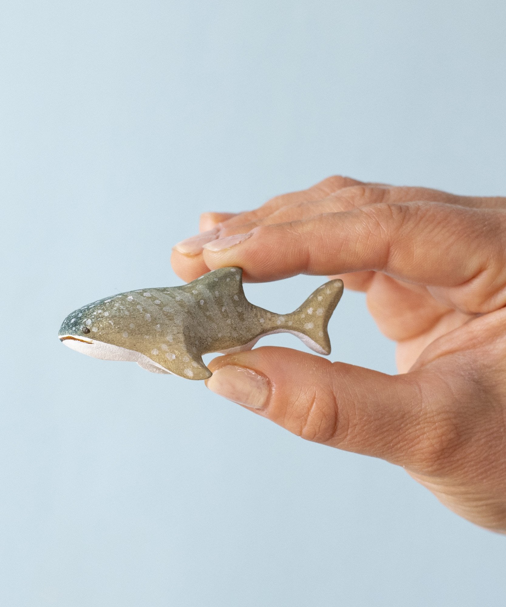 An adult hand holding the Bumbu handmade and hand painted wooden grey baby whale shark figure showing scale