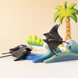 Bumbu handmade and hand painted wooden black manta ray figure available at Babipur and resting on a wooden wave piece with palm trees in the background.