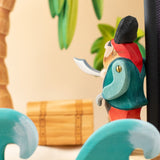 Bumbu handmade and hand painted pirate wooden figure in a ocean play scene looking towards a treasure chest under a palm tree with ocean waves in front