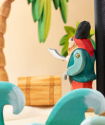 Bumbu handmade and hand painted pirate wooden figure in a ocean play scene looking towards a treasure chest under a palm tree with ocean waves in front