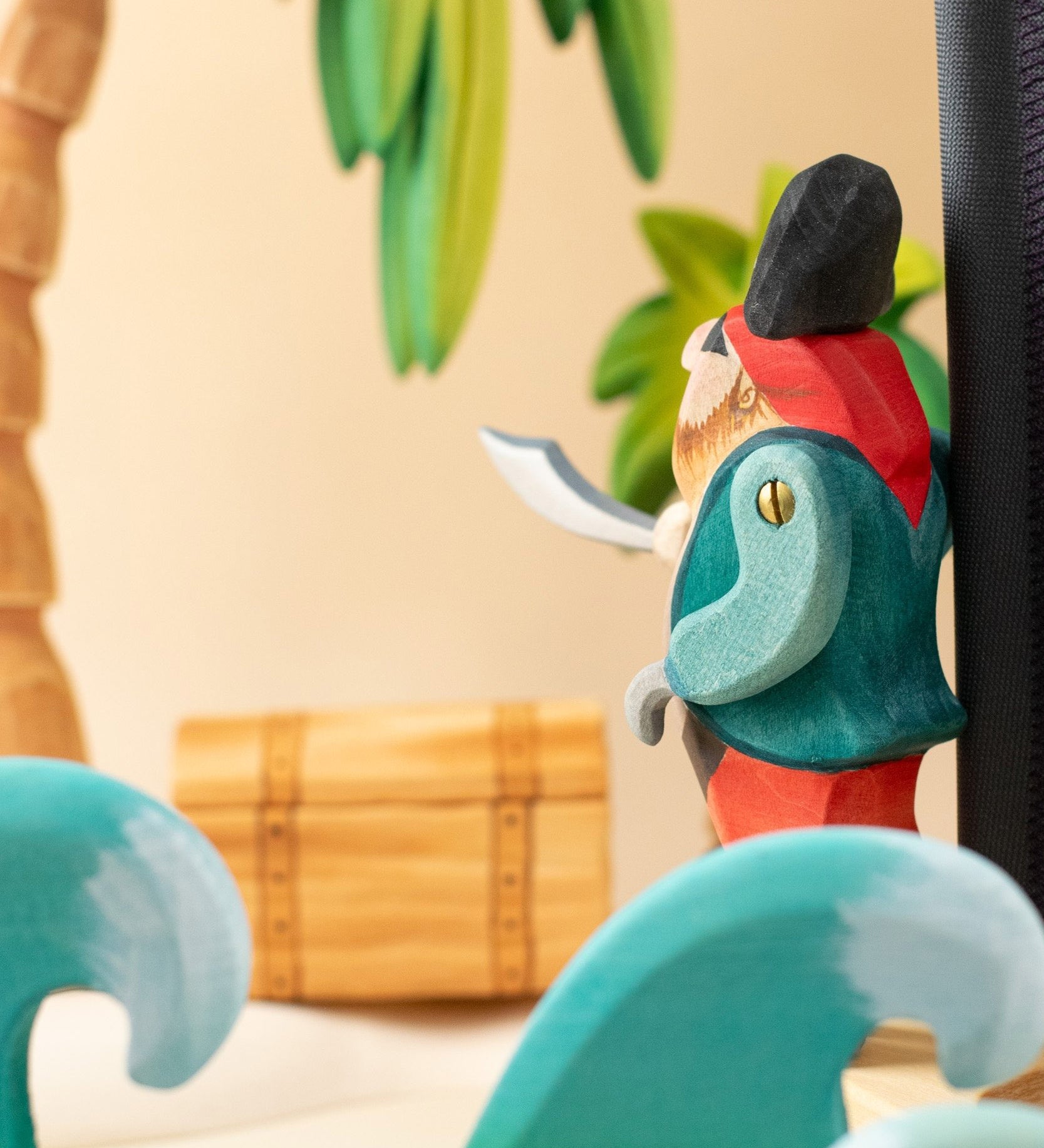 Bumbu handmade and hand painted pirate wooden figure in a ocean play scene looking towards a treasure chest under a palm tree with ocean waves in front