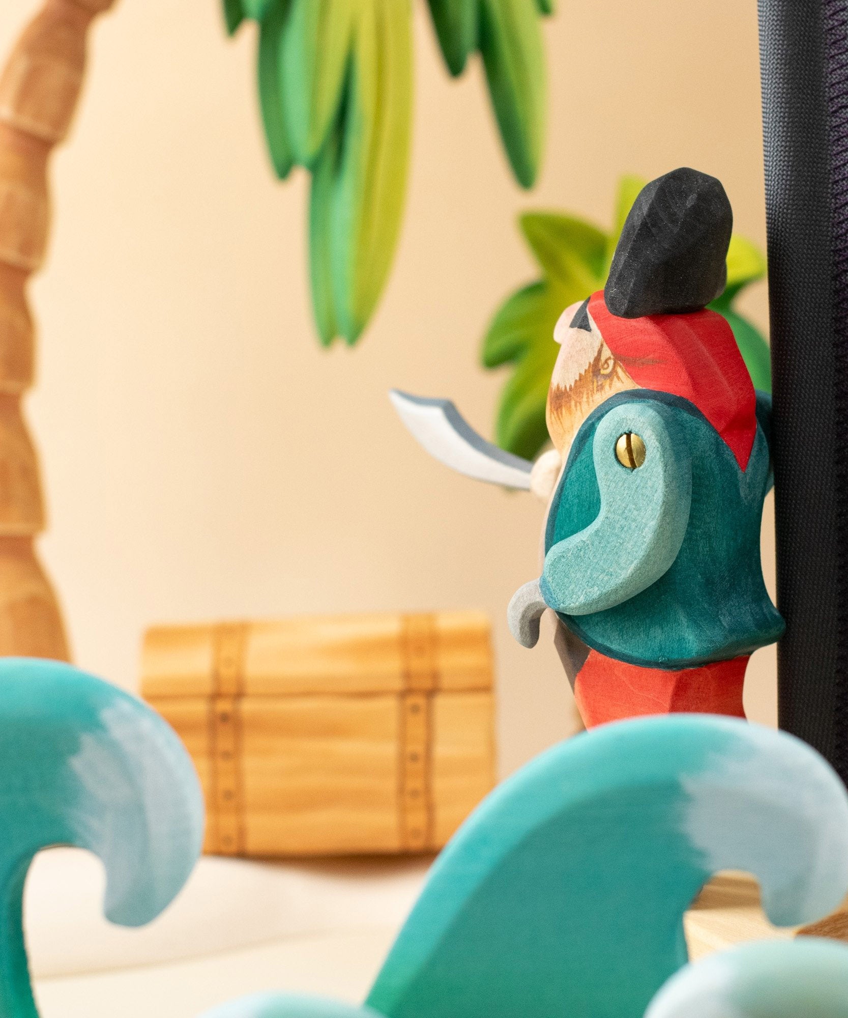Bumbu handmade and hand painted pirate wooden figure in a ocean play scene looking towards a treasure chest under a palm tree with ocean waves in front