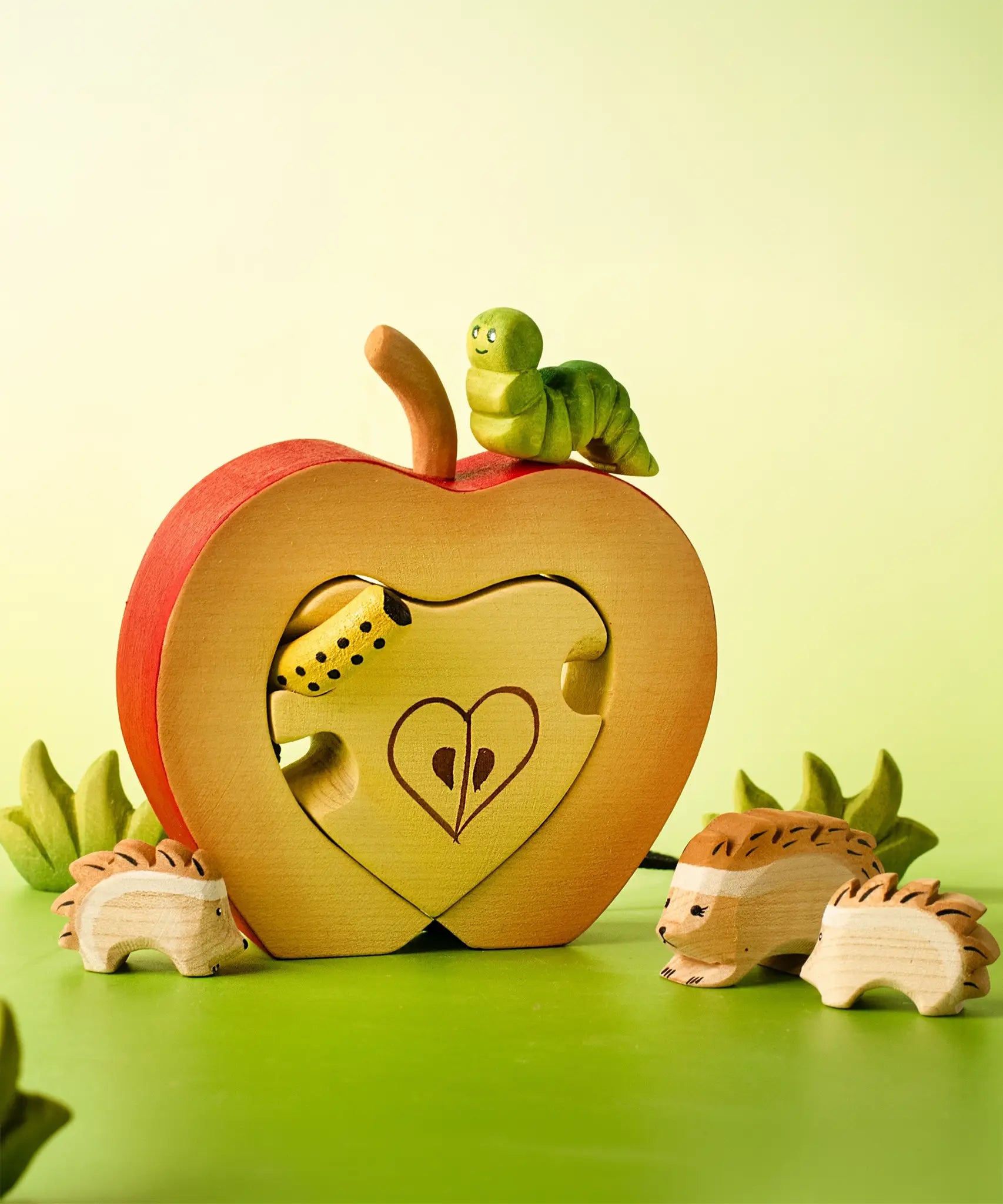 Bumbu handcrafted wooden green caterpillar figure standing on a apple puzzle set with a hedgehog family figures in front