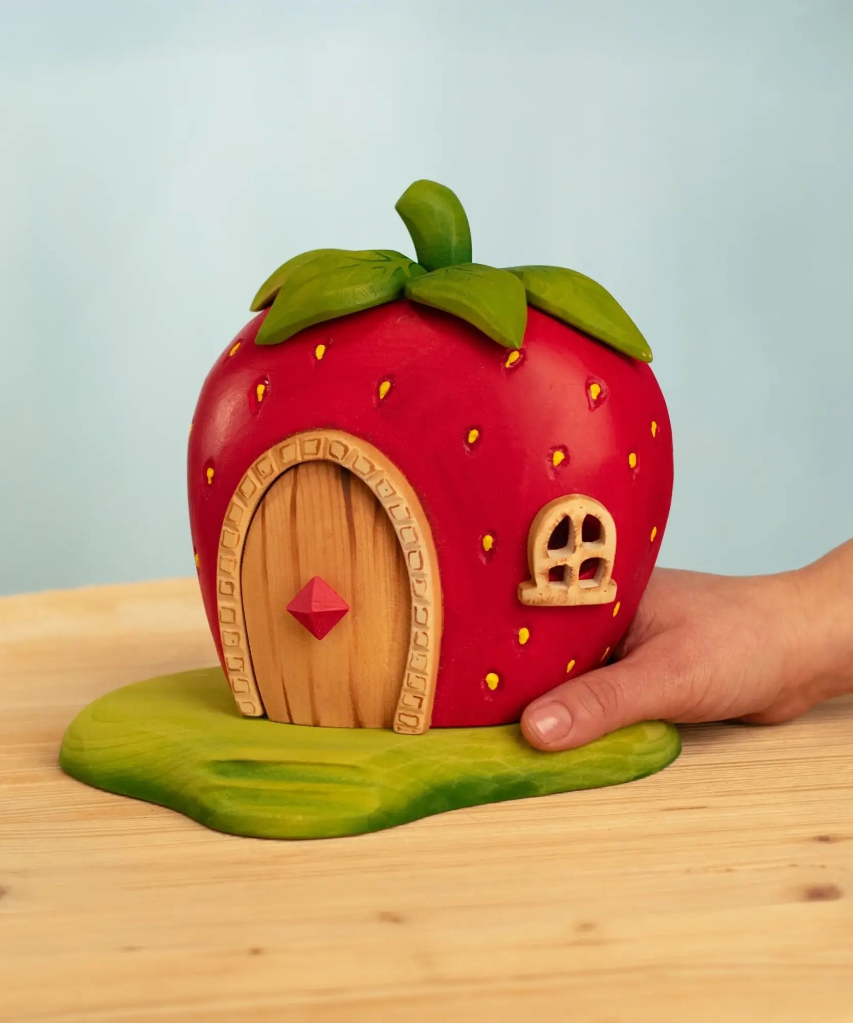 Adult hand holding Bumbu wooden strawberry house standing on a green base 