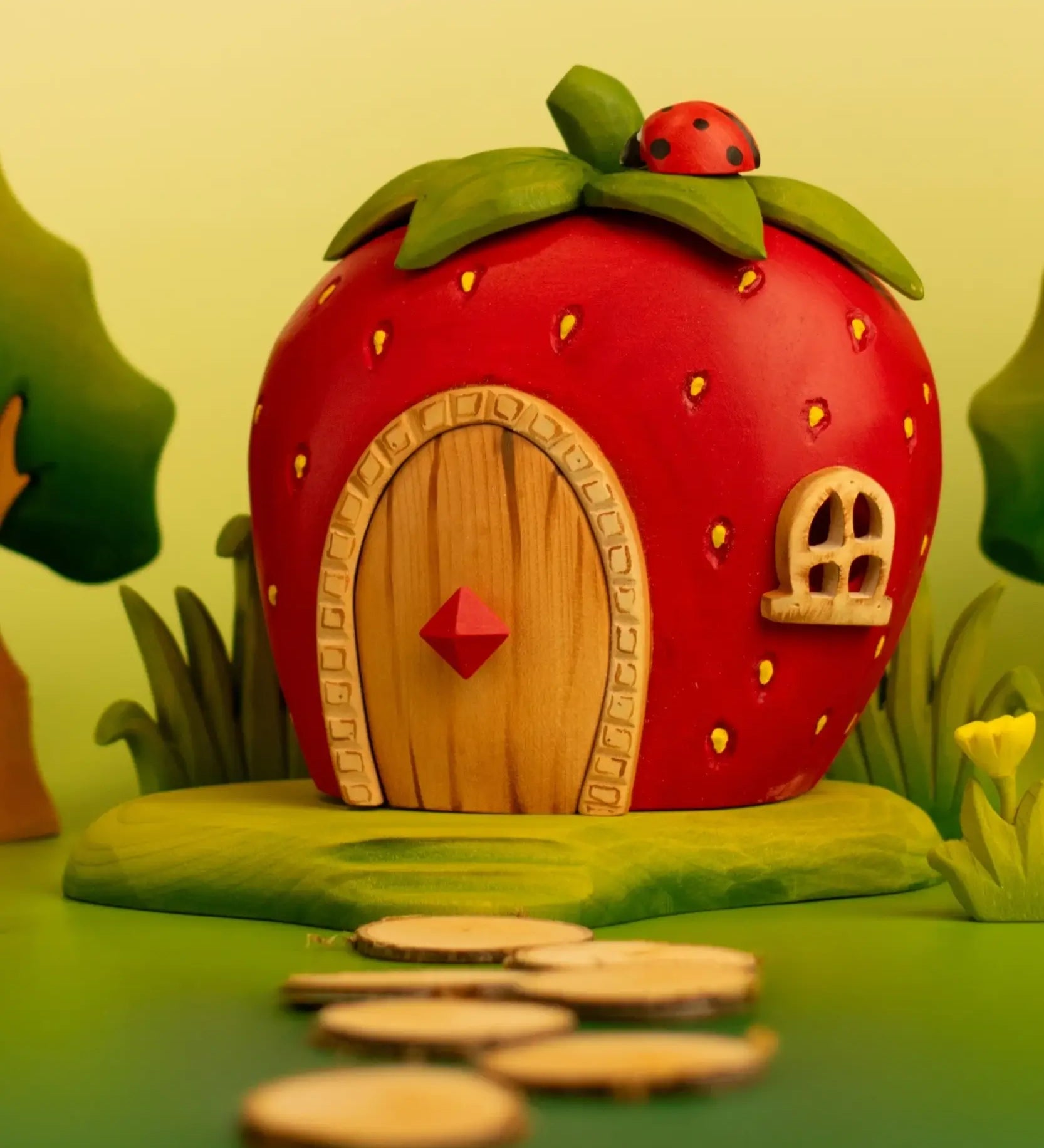 Bumbu wooden strawberry house standing on a green base and ladybird on top in a playscene with trees, grass and flowers
