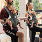 Two people working out and exercising at home, both carrying a child in a Carifit+ Baby Carrier in Cool Grey. The child on the left is outwards facing in the carrier, and the child on the right is inwards facing. The image shows how supported the child is inside the carrier and that parents can still exercise whist carrying their children in the carrier