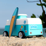 The back of the Candylab VW Beach bus in blue, with a blue and surf board propped up next to the van, stood on a sandy beach with a blue sky background. The image also shows colourful stickers on the back of the van