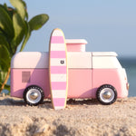 A side view of the Candylab VW Beach bus in pink, with a pink and white stripe surf board propped up next to the van, stood on a sandy beach with a blue sky background