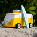 Candylab VW Beach bus in yellow, with a blue surf board propped up next to the van, stood on a sandy beach with green tree leaves and branches in the background
