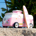 Candylab VW Beach bus in pink, with a pink and white stripe surf board propped up next to the van, stood on a sandy beach with green tree leaves and branches in the background