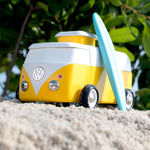 Candylab VW Beach bus in yellow, with a blue surf board propped up next to the van, stood on a sandy beach with green tree leaves and branches in the background. The image shows the front of the bus with a VW logo and silver studs to mimic head-lights